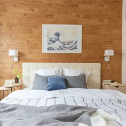 Sypialnia z drewnianą ścianą, oświetleniem punktowym i łóżkiem z tapicerowanym zagłówkiem, nad którym wisi obraz przedstawiający falę.