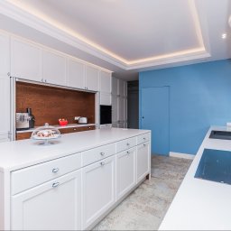 Wasze Wnętrze - Nowoczesna kuchnia z białymi szafkami i wyspą, drewnianym akcentem na ścianie i niebieską ścianą w tle. Na blacie ekspres do kawy i ciasto pod kloszem.