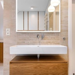 Wasze Wnętrze - Nowoczesna łazienka z drewnianą szafką pod umywalką, prostokątną białą umywalką i podświetlanym lustrem. Beżowe płytki na ścianach i podłodze. Minimalistyczne oświetlenie.