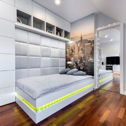 Wasze Wnętrze - Pokój młodzieżowy z łóżkiem na podeście z podświetleniem LED, tapicerowanym wezgłowiem i szafkami w zabudowie. Na ścianie fototapeta z panoramą miasta.