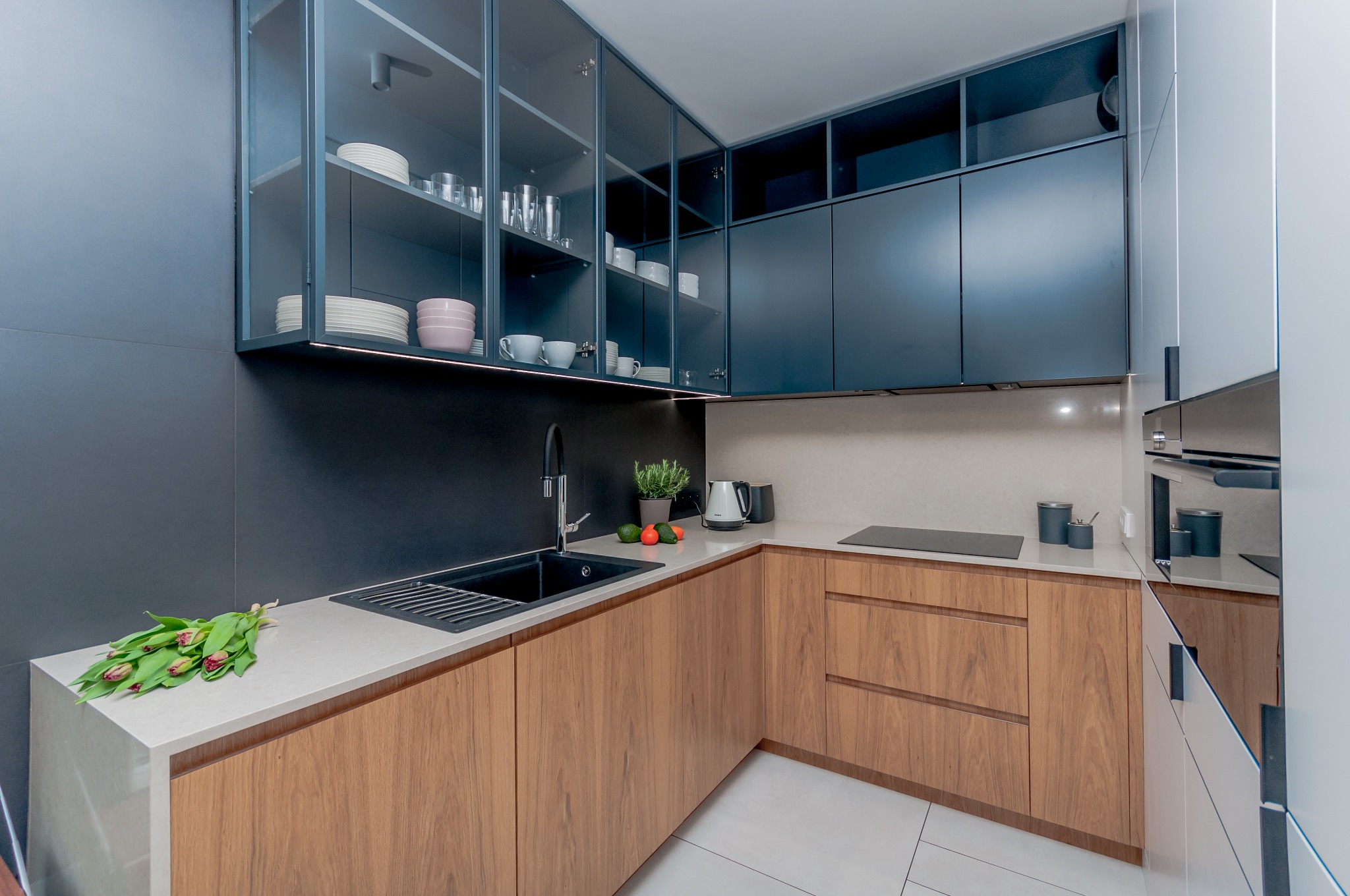 Nowoczesna kuchnia z drewnianymi frontami, czarnym zlewem i przeszklonymi szafkami wiszącymi. Na blacie kwiaty i akcesoria kuchenne. Minimalistyczny design.