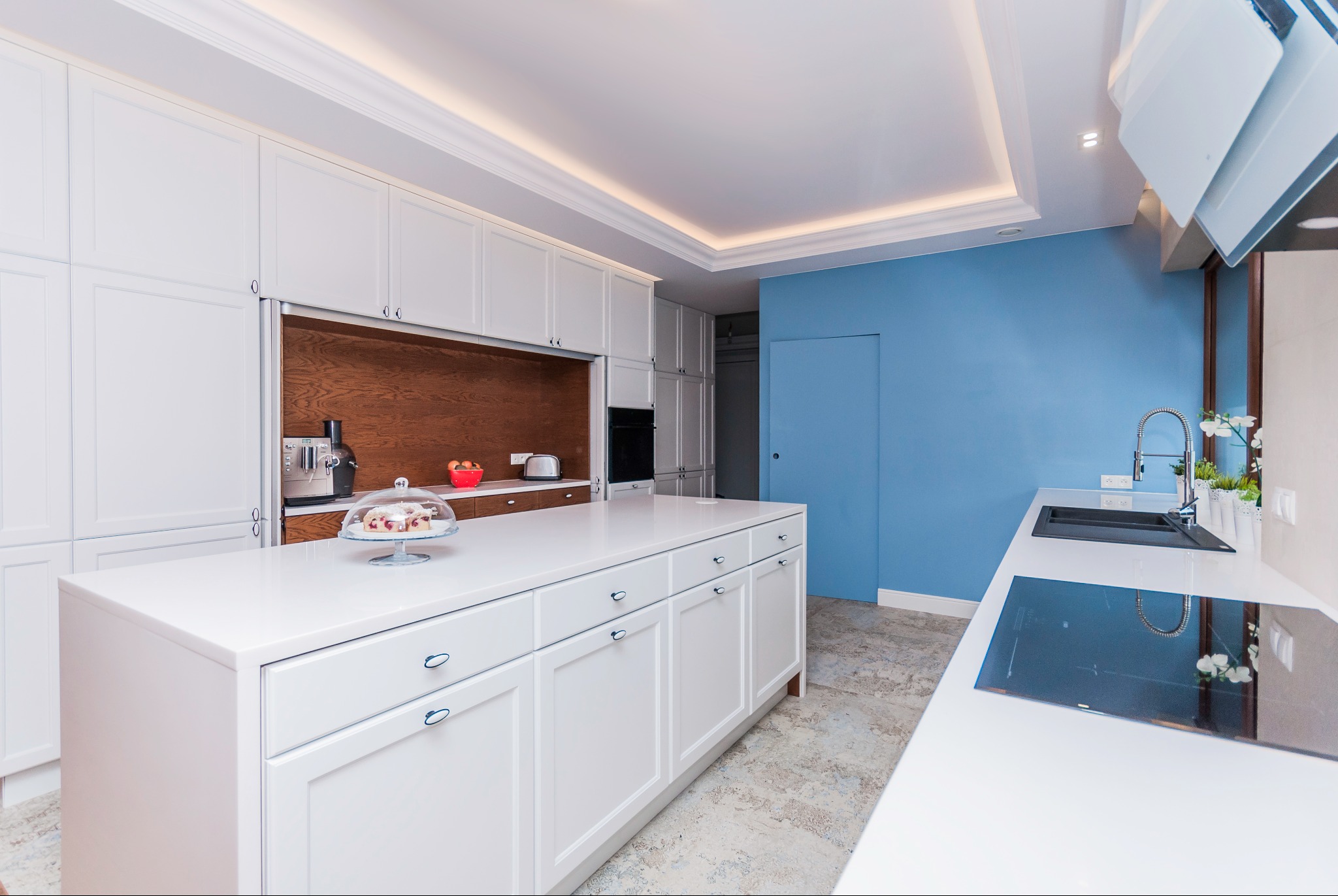 Nowoczesna kuchnia z białymi szafkami i wyspą, drewnianym akcentem na ścianie i niebieską ścianą w tle. Na blacie ekspres do kawy i ciasto pod kloszem.