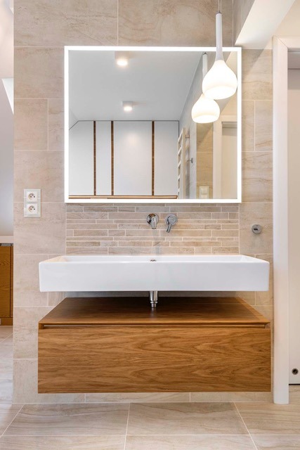 Nowoczesna łazienka z drewnianą szafką pod umywalką, prostokątną białą umywalką i podświetlanym lustrem. Beżowe płytki na ścianach i podłodze. Minimalistyczne oświetlenie.