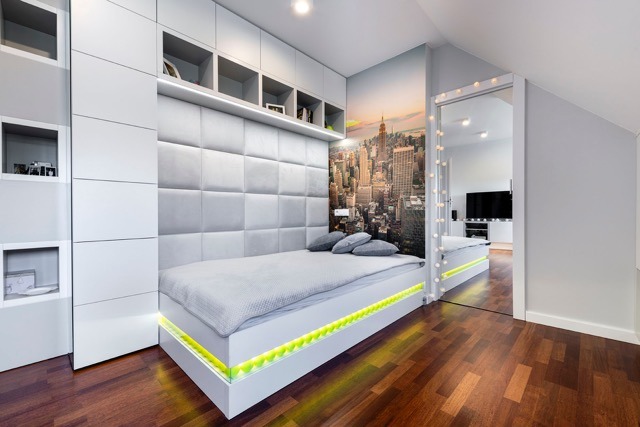 Pokój młodzieżowy z łóżkiem na podeście z podświetleniem LED, tapicerowanym wezgłowiem i szafkami w zabudowie. Na ścianie fototapeta z panoramą miasta.