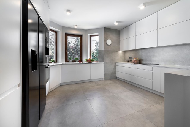 Nowoczesna, jasna kuchnia z białymi szafkami bez uchwytów i czarną lodówką. Okno z widokiem na zieleń. Minimalistyczny design z szarym blatem i płytkami.