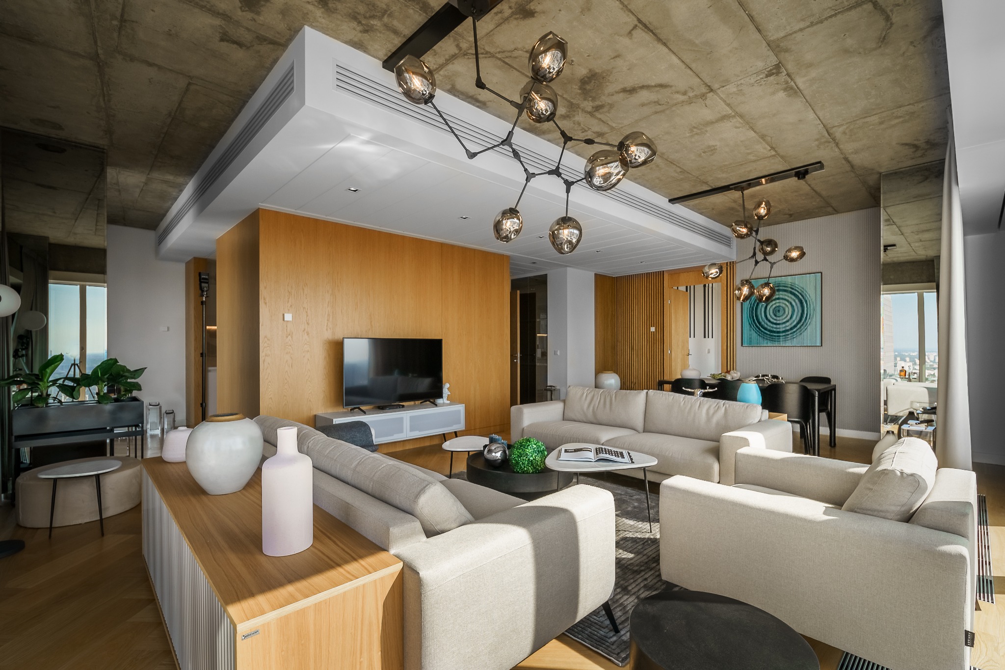 Nowoczesny salon w Płochocinie z drewnianą zabudową TV, beżowymi sofami i betonowym sufitem. Designerska lampa sufitowa dodaje wnętrzu charakteru.