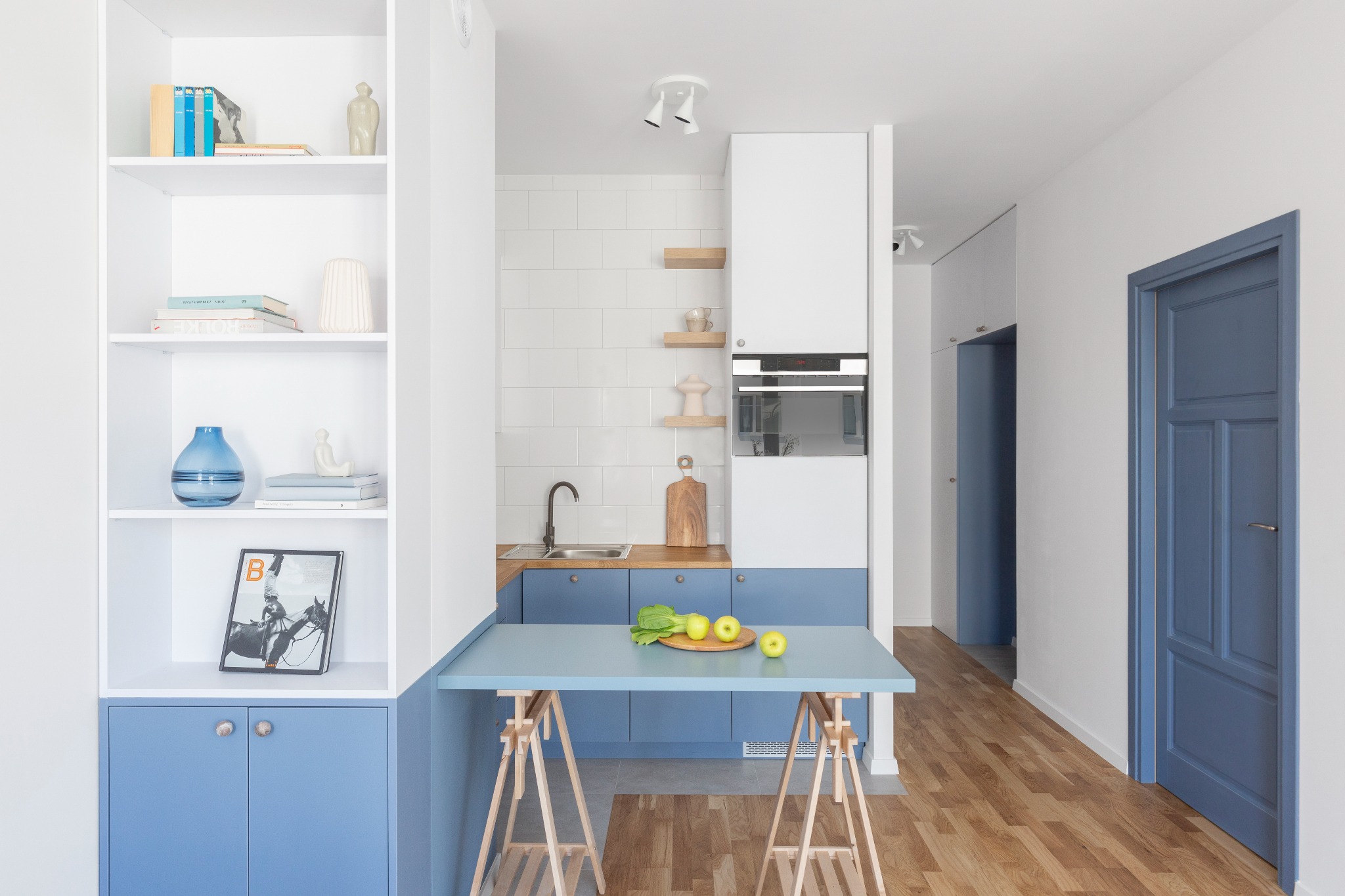 Jasne wnętrze kuchni z niebieskimi akcentami: szafki, blat na kozłach, półki z dekoracjami. Minimalistyczny design, drewniana podłoga i drzwi w kolorze niebieskim.