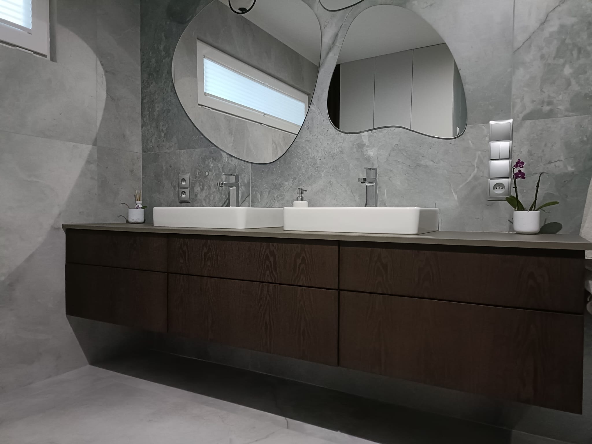 Nowoczesna łazienka z podwójną umywalką nablatową, szafką z ciemnego drewna i asymetrycznymi lustrami na tle szarej ściany z efektem kamienia.
