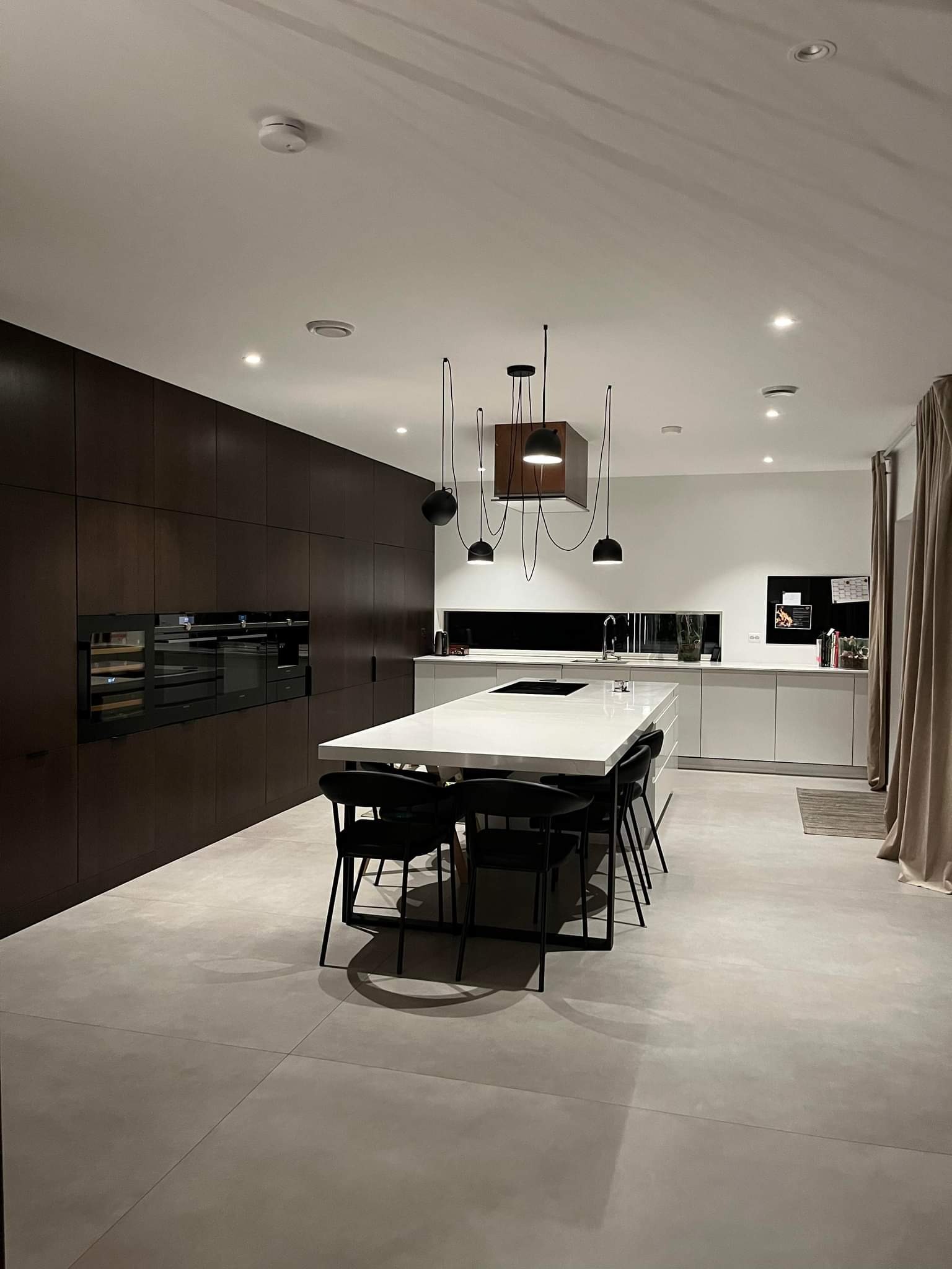 Nowoczesna kuchnia z wyspą i ciemnymi szafkami. Minimalistyczne lampy wiszące nad blatem. W tle okno i jasne szafki. Widoczny sprzęt AGD w zabudowie.