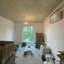Paweł Kaczówka CONSULTING - Pokój w trakcie prac wykończeniowych: surowy strop, ściany po wstępnym malowaniu, okno z widokiem na zieleń, materiały budowlane i narzędzia porozrzucane na podłodze.
