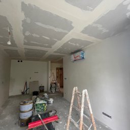 Paweł Kaczówka CONSULTING - Wnętrze w trakcie prac wykończeniowych: surowy sufit z widocznymi łączeniami płyt kartonowo-gipsowych i niedoskonałościami, ściany po nałożeniu gładzi, drabina malarska, wiadro z farbą i narzędzia...