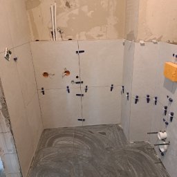 Paweł Kaczówka CONSULTING - Świeżo położone szare płytki podłogowe i jasne płytki ścienne w trakcie montażu z widocznymi krzyżykami dystansowymi, otwory na instalacje elektryczne i wodne, surowe ściany w tle.