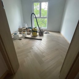 Paweł Kaczówka CONSULTING - Pokój po malowaniu ścian na szaro, z parkietem ułożonym w jodełkę, widoczne pędzle, wałki, kuwety malarskie i odkurzacz przemysłowy przy oknie.