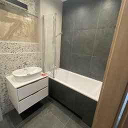 Paweł Kaczówka CONSULTING - Nowoczesna łazienka z umywalką nablatową na szafce z dwiema szufladami, kabiną prysznicową nad wanną, szarą glazurą i fragmentem ściany z dekoracyjnym tynkiem mozaikowym.
