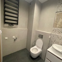 Paweł Kaczówka CONSULTING - Nowoczesna łazienka z szarą płytką, wiszącą toaletą i grzejnikiem drabinkowym w kolorze czarnym, uzupełniona umywalką nablatową z szafką i fragmentem ściany z mozaiką.