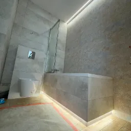 Nowoczesna łazienka z podświetlaną wanną obudowaną płytkami, wiszącą toaletą z chromowanym przyciskiem spłukiwania i szklanym parawanem nawannowym, podłoga zabezpieczona folią malarską.