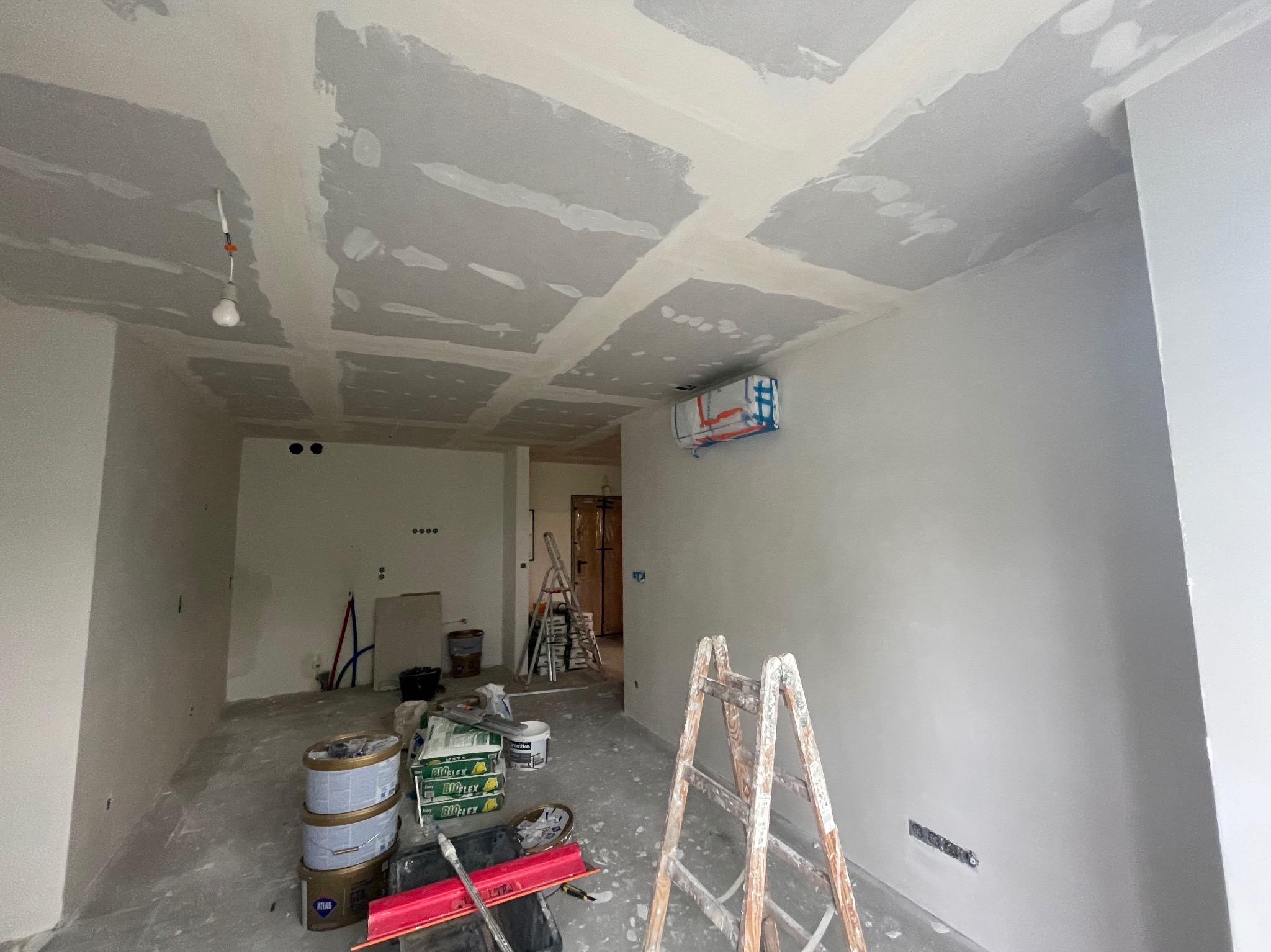 Wnętrze w trakcie prac wykończeniowych: surowy sufit z widocznymi łączeniami płyt kartonowo-gipsowych i niedoskonałościami, ściany po nałożeniu gładzi, drabina malarska, wiadro z farbą i narzędzia...