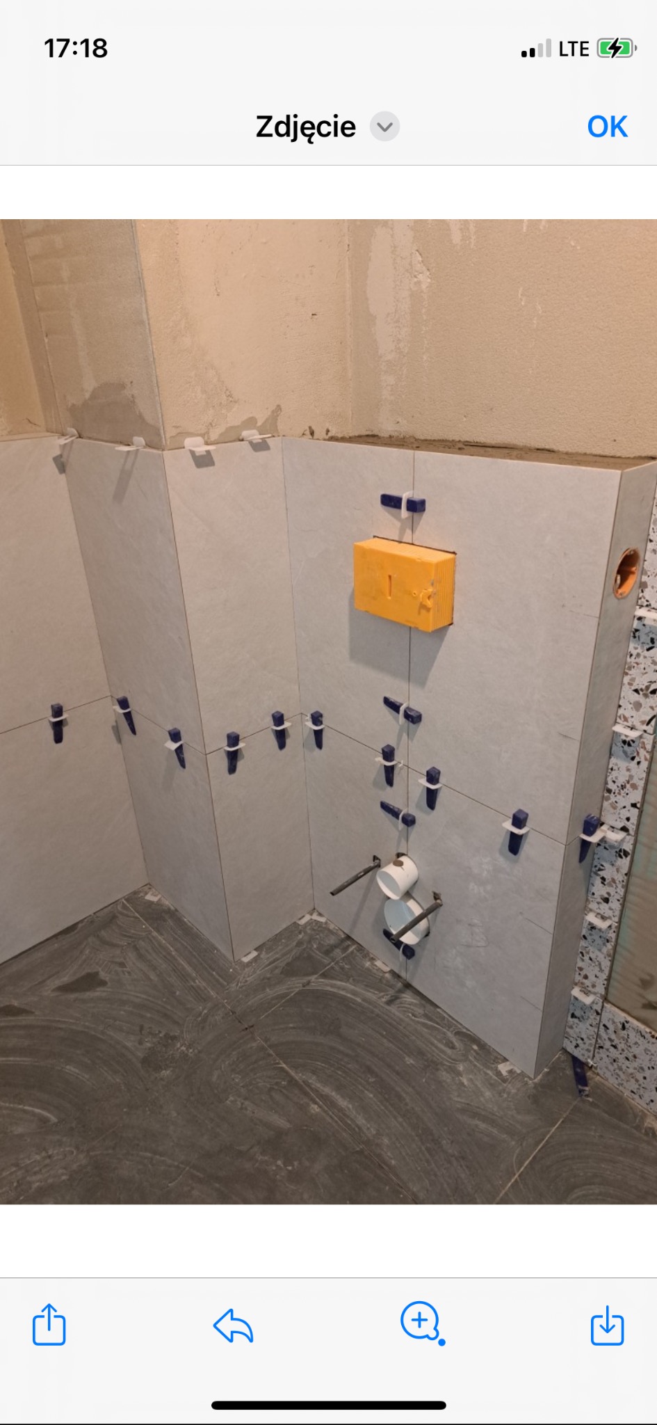 Świeżo ułożone, jasnoszare płytki ścienne w łazience, zabezpieczone niebieskimi krzyżykami dystansowymi, z widocznymi rurami i żółtą poziomicą w narożniku.