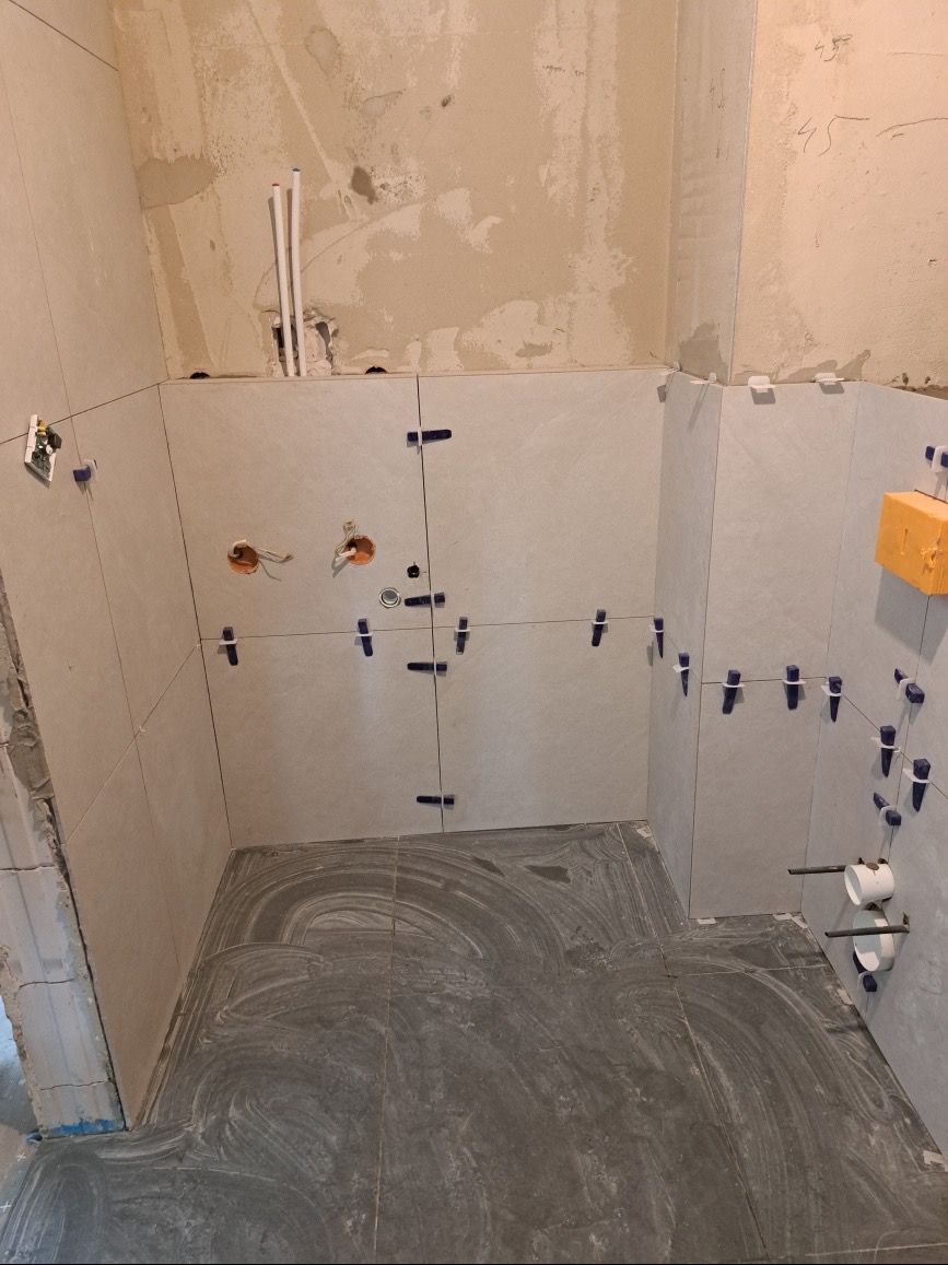 Świeżo położone szare płytki podłogowe i jasne płytki ścienne w trakcie montażu z widocznymi krzyżykami dystansowymi, otwory na instalacje elektryczne i wodne, surowe ściany w tle.