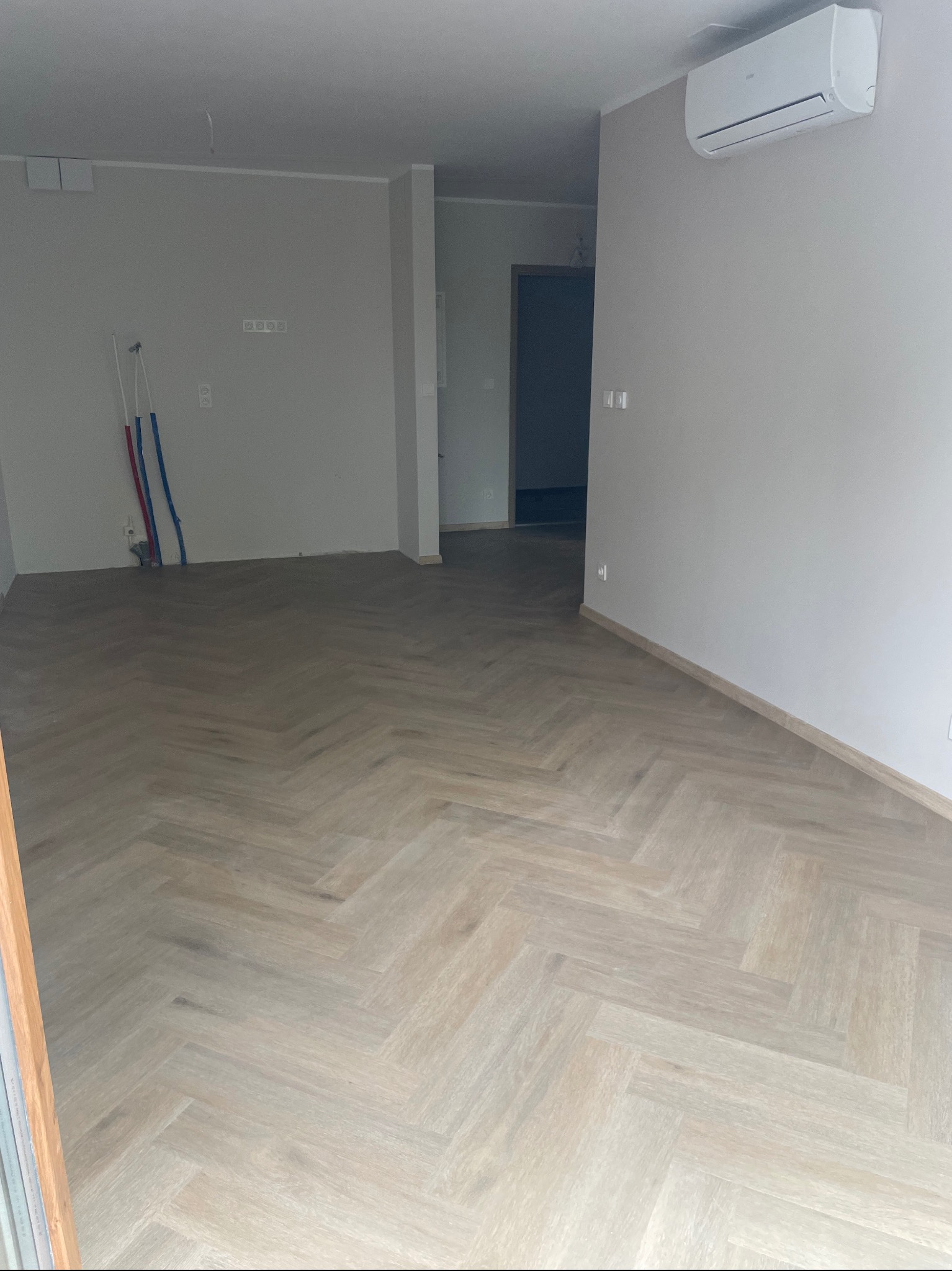 Pusty pokój z jasną podłogą ułożoną w jodełkę, rury wystające ze ściany i klimatyzator na ścianie, widok z poziomu podłogi.
