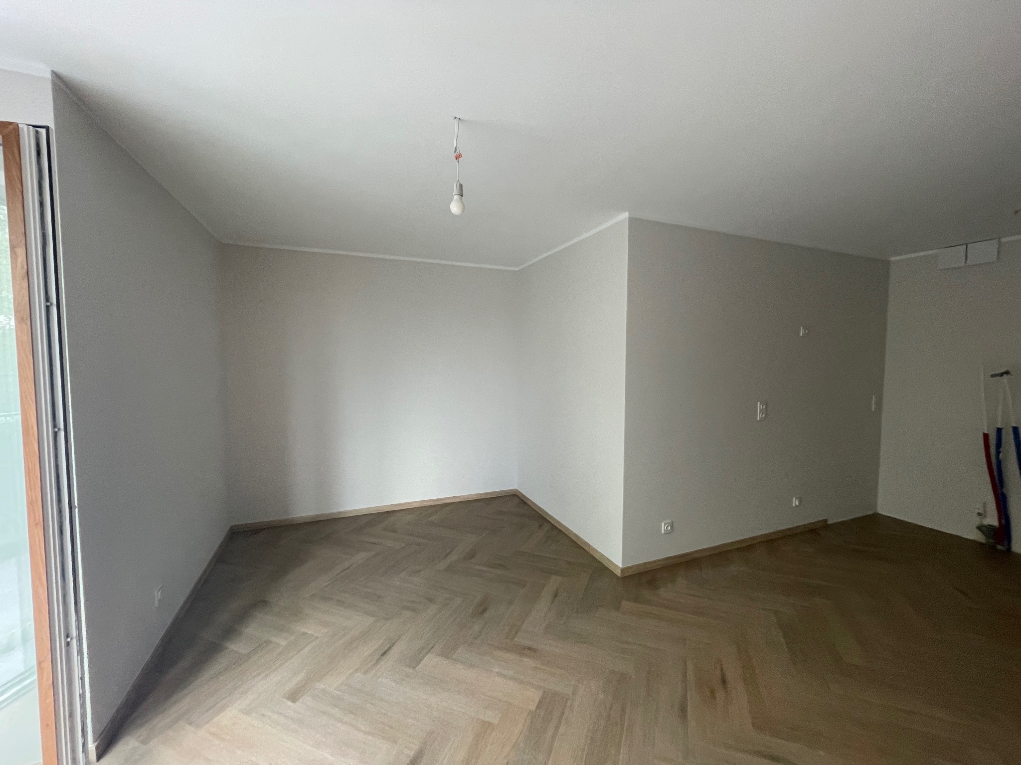 Pusty pokój z parkietem ułożonym w jodełkę, szare ściany, pojedyncza żarówka zwisająca z sufitu, widoczne gniazdka elektryczne i rury przy ścianie.