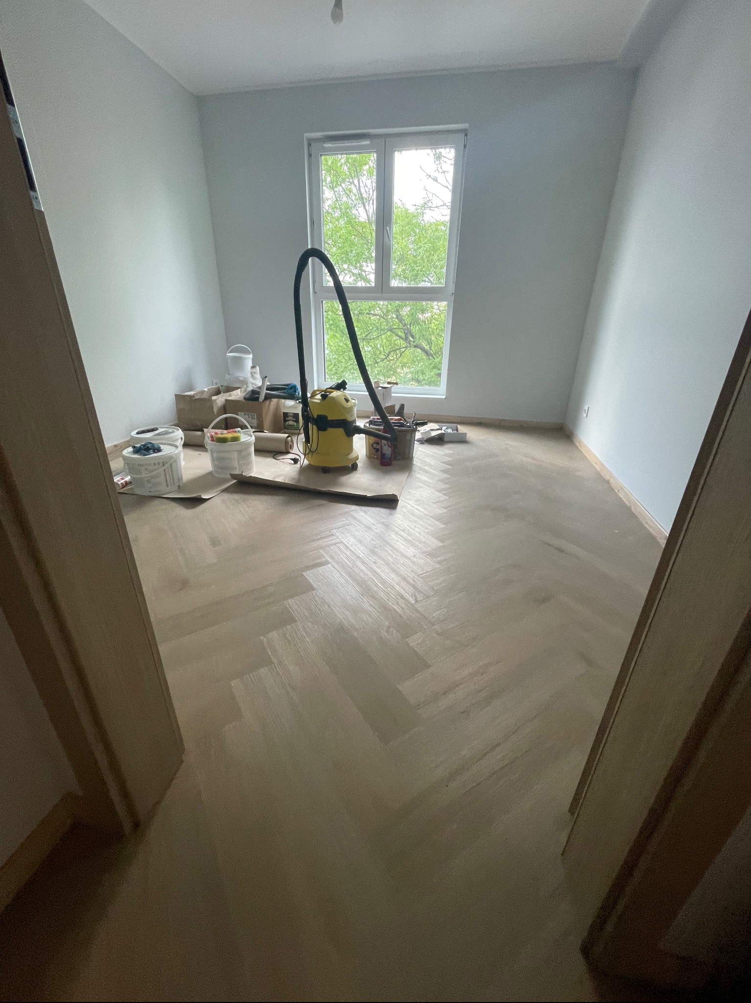 Pokój po malowaniu ścian na szaro, z parkietem ułożonym w jodełkę, widoczne pędzle, wałki, kuwety malarskie i odkurzacz przemysłowy przy oknie.