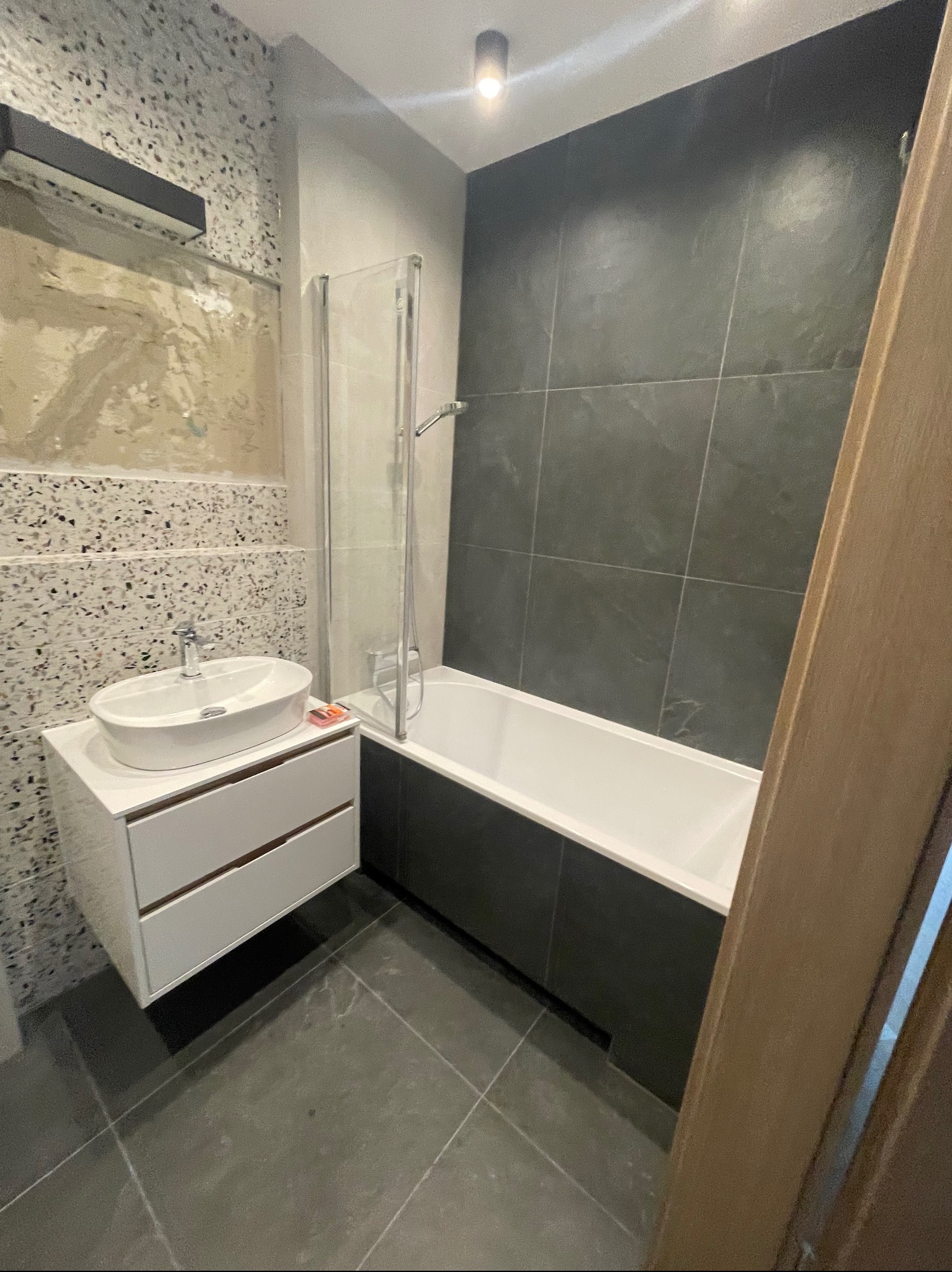 Nowoczesna łazienka z umywalką nablatową na szafce z dwiema szufladami, kabiną prysznicową nad wanną, szarą glazurą i fragmentem ściany z dekoracyjnym tynkiem mozaikowym.