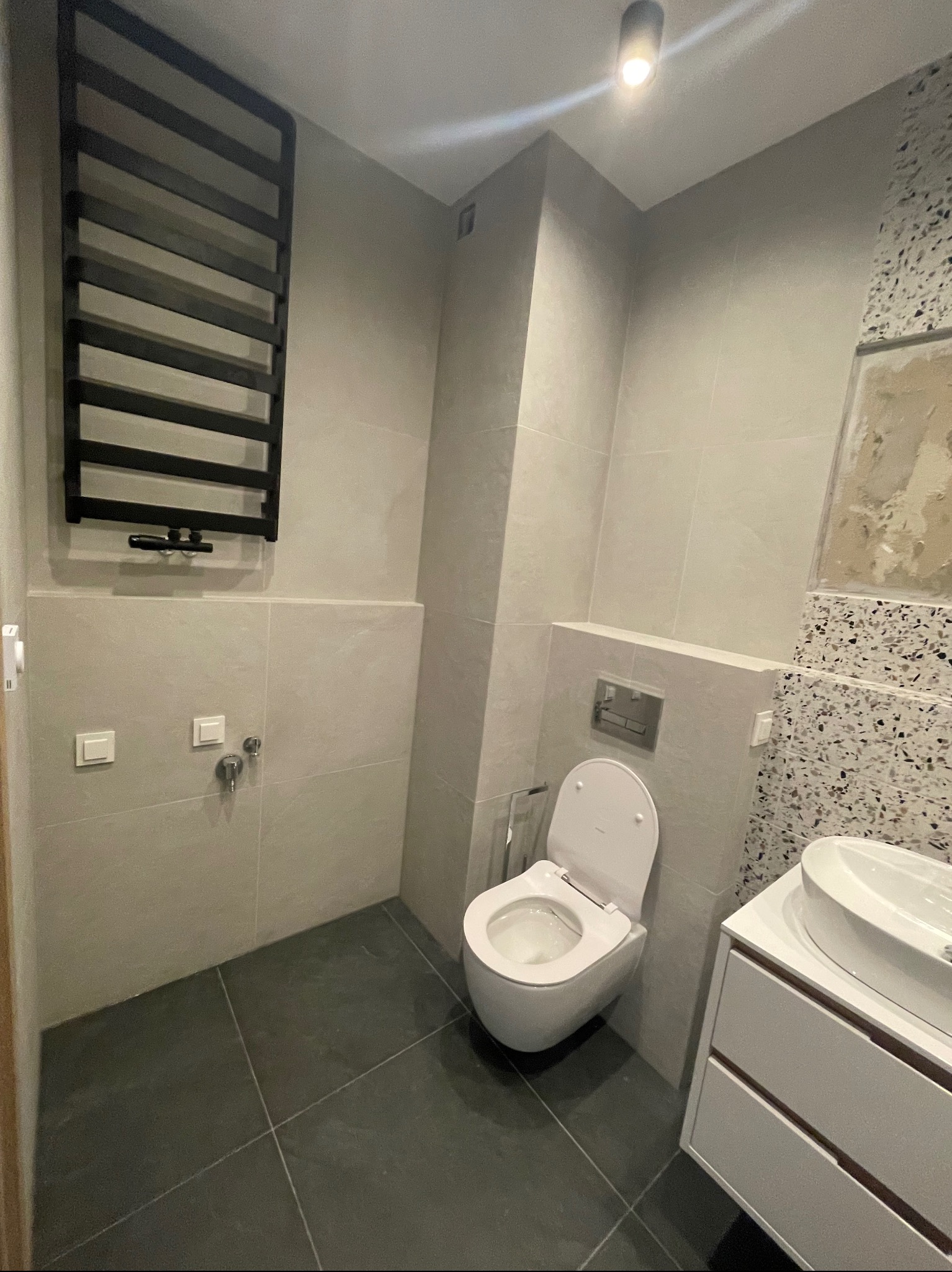 Nowoczesna łazienka z szarą płytką, wiszącą toaletą i grzejnikiem drabinkowym w kolorze czarnym, uzupełniona umywalką nablatową z szafką i fragmentem ściany z mozaiką.