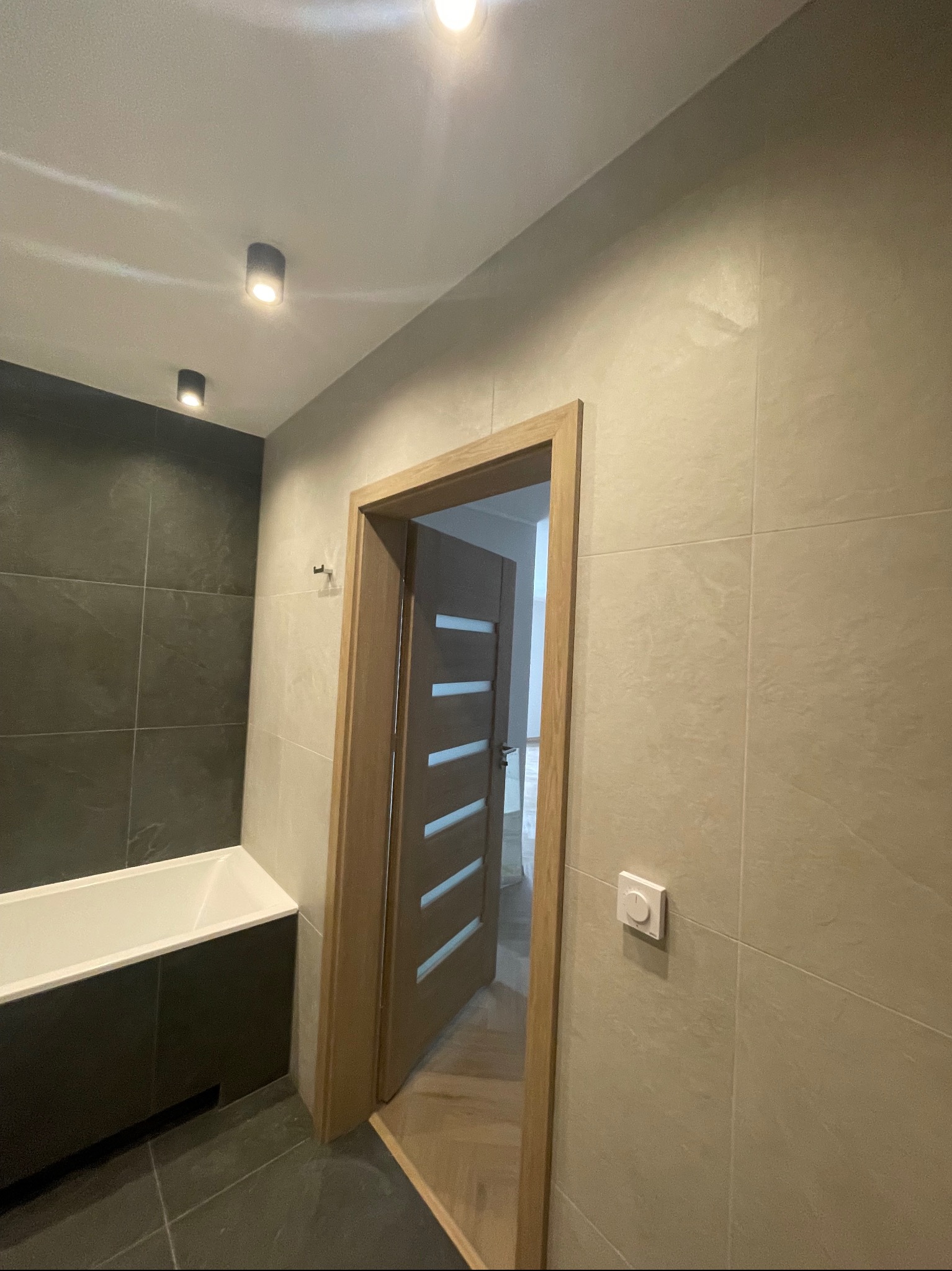 Wykończona łazienka z czarną wanną, jasnymi płytkami na ścianach, drewnianą ościeżnicą i drzwiami z poziomymi przeszkleniami oraz punktowym oświetleniem sufitowym.