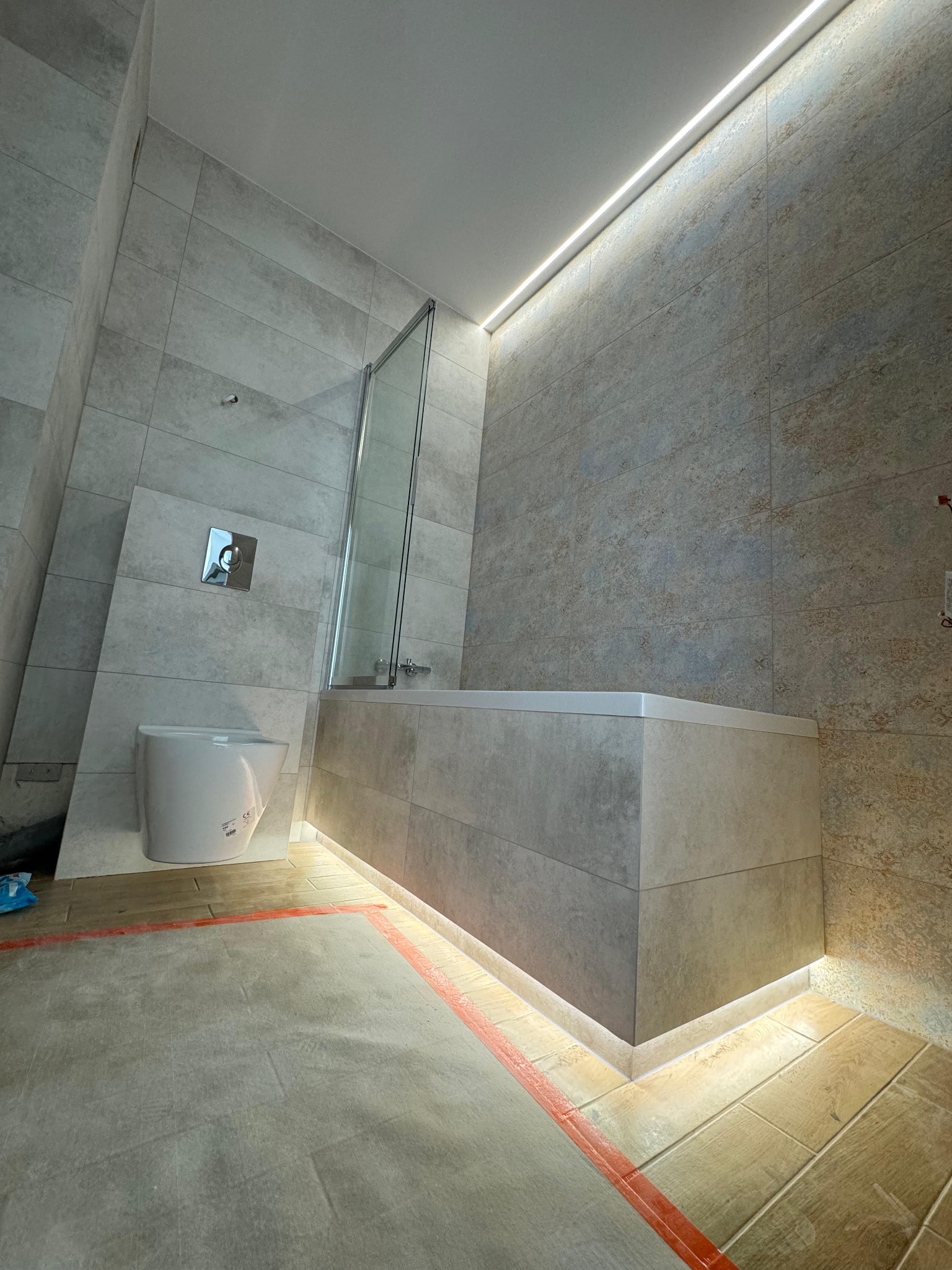 Nowoczesna łazienka z podświetlaną wanną obudowaną płytkami, wiszącą toaletą z chromowanym przyciskiem spłukiwania i szklanym parawanem nawannowym, podłoga zabezpieczona folią malarską.