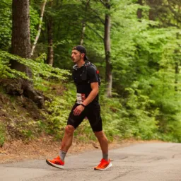 Mężczyzna w czarnym stroju sportowym i pomarańczowych butach biegnie pod górę asfaltową drogą, otoczony zielonym lasem, z numerem startowym 3009 przypiętym do koszulki.