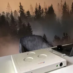 Aparat TimeWaver na białym biurku z tapetą leśną w tle, obok laptop i myszka.