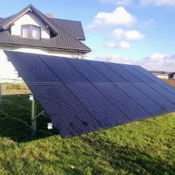 Instalacja fotowoltaiczna na trawniku w Olsztynie, panele skierowane w stronę słońca, w tle dom jednorodzinny z szarą dachówką i błękitne niebo z chmurami.