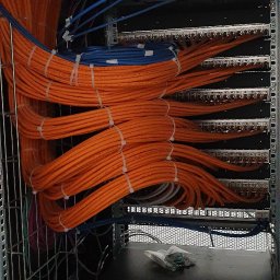 Is Tech Igor Szablewski - Zorganizowana wiązka pomarańczowych i niebieskich kabli sieciowych przymocowanych opaskami zaciskowymi w szafie rack, z widocznymi panelami krosowniczymi i przełącznikami sieciowymi.