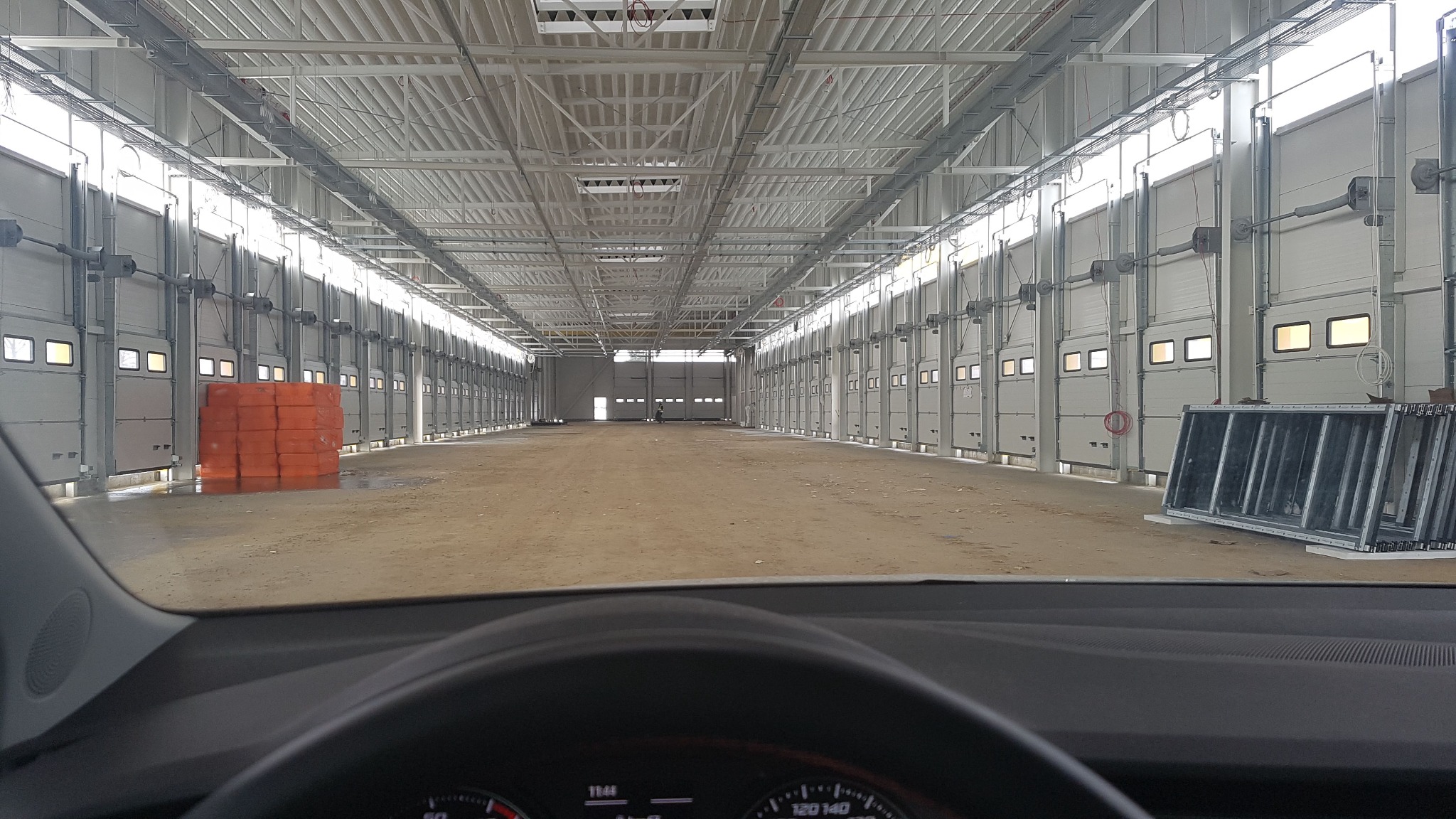 Wnętrze hali magazynowej w trakcie budowy z widokiem z wnętrza samochodu, widoczne liczne bramy garażowe, konstrukcja dachu i materiały budowlane.