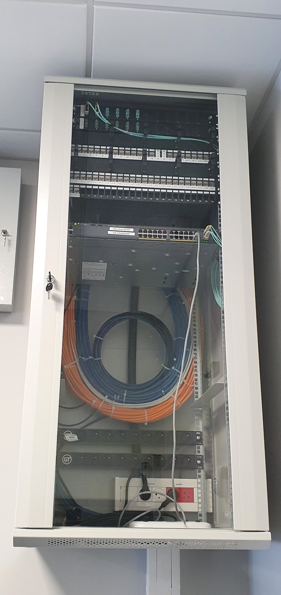 Szafa rack TOTEN z transparentnymi drzwiami, zawierająca patch panel, switch sieciowy, oraz uporządkowane kable Ethernet w kolorach pomarańczowym, białym i niebieskim, zasilanie awaryjne na dole.