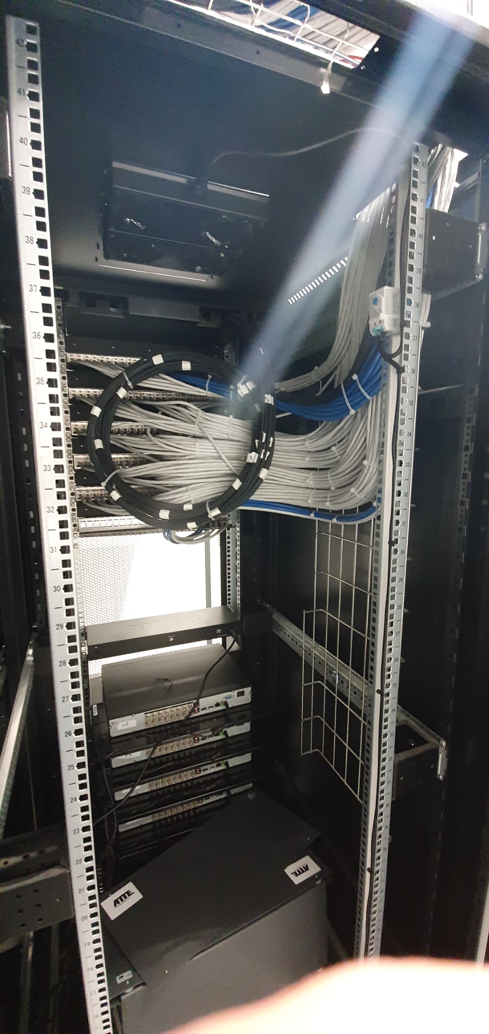 Wnętrze szafy serwerowej z uporządkowanymi kablami w kolorach białym, szarym i niebieskim, ułożonymi w okrągłe wiązki, widoczne urządzenia sieciowe na półkach, oraz metalowa konstrukcja szafy...