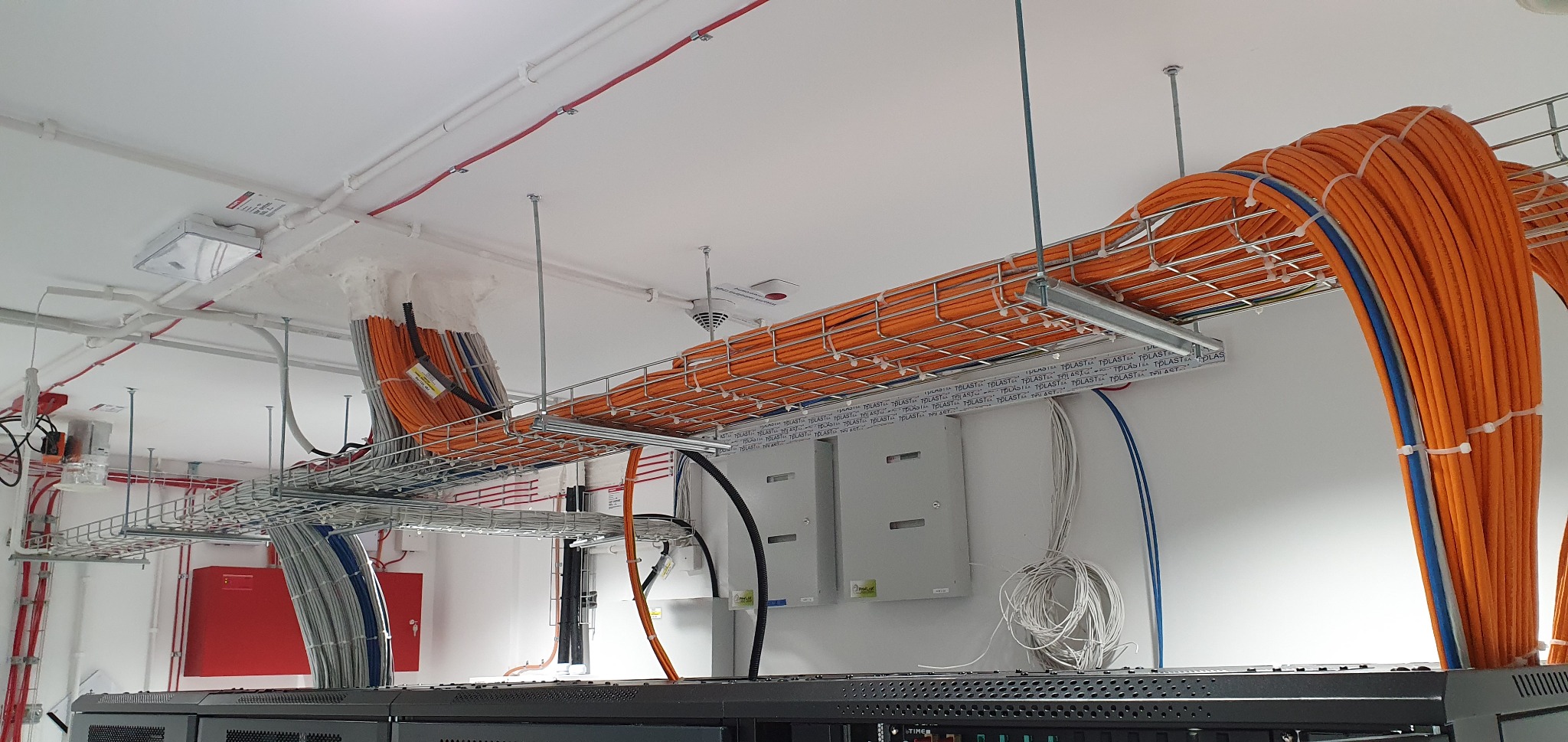 Widok na system koryt kablowych pod sufitem, wypełnionych pomarańczowymi przewodami z niebieskimi akcentami, mocowanych za pomocą białych opasek zaciskowych, z widocznymi białymi puszkami...