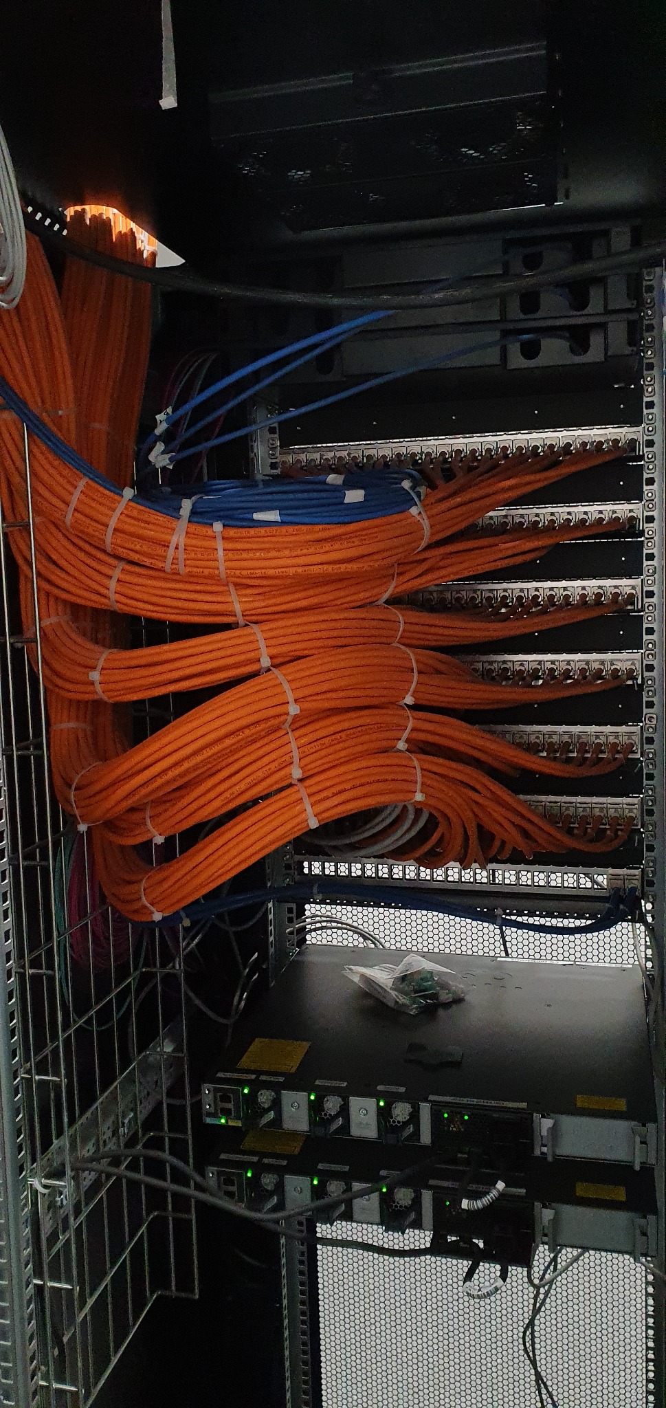Zorganizowana wiązka pomarańczowych i niebieskich kabli sieciowych przymocowanych opaskami zaciskowymi w szafie rack, z widocznymi panelami krosowniczymi i przełącznikami sieciowymi.