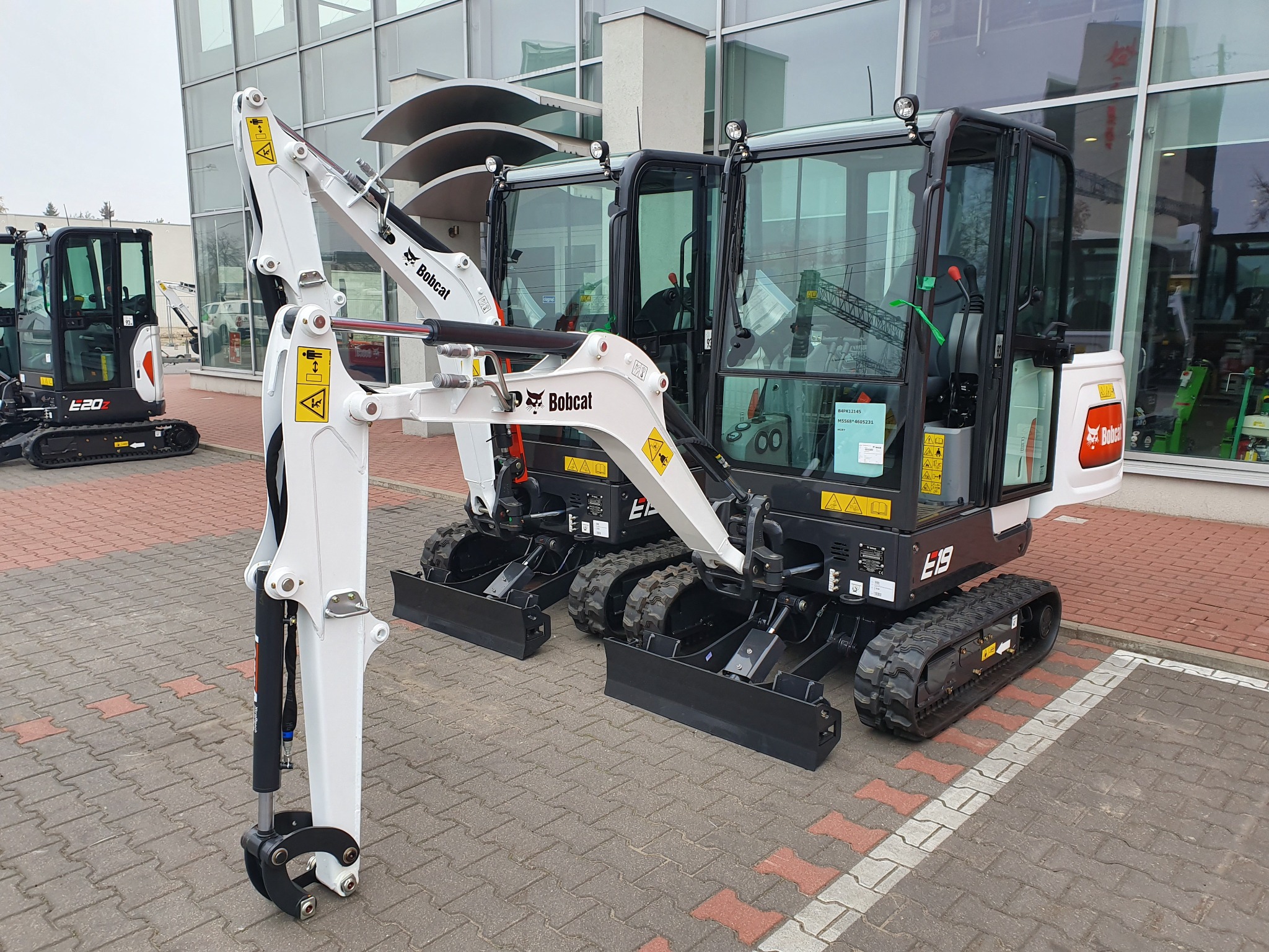Dwa nowe mini-koparki Bobcat E19 ustawione obok siebie na kostce brukowej, z widocznym ramieniem kopiącym i kabiną operatora, na tle przeszklonego budynku.