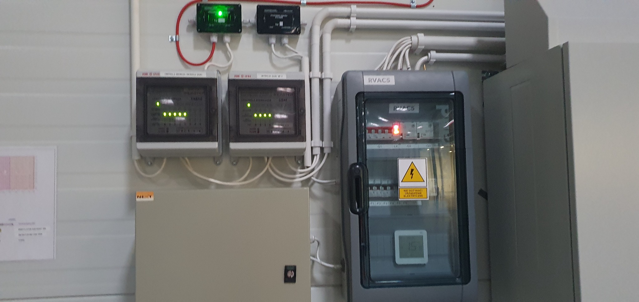 Panel sterowania systemem alarmowym z sygnalizacją LED i tabliczką ostrzegawczą, widoczne przewody i rury instalacyjne.