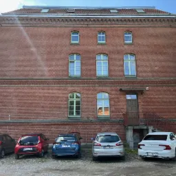 Ceglany budynek biurowy z zaparkowanymi samochodami, widoczny znak z numerem 8 na ścianie.