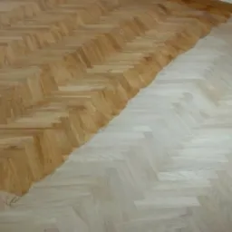 Fragment podłogi z parkietu ułożonego w jodełkę, częściowo po procesie lakierowania lub olejowania, z widocznym kontrastem między surowym, jasnym drewnem a zabezpieczoną, ciemniejszą powierzchnią.