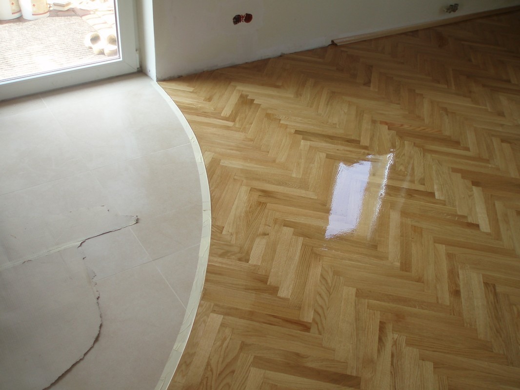 Świeżo polakierowany parkiet ułożony w jodełkę, przechodzący w jasną, nierówną posadzkę przy balkonie, widoczne odbicie światła w lakierze.