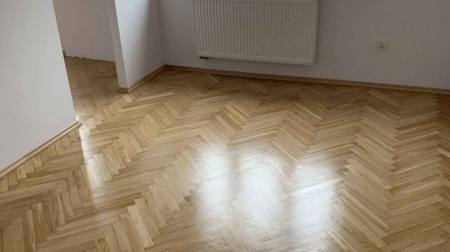 Jasny parkiet ułożony we wzór jodełki francuskiej w odnowionym pokoju z białymi ścianami i grzejnikiem.