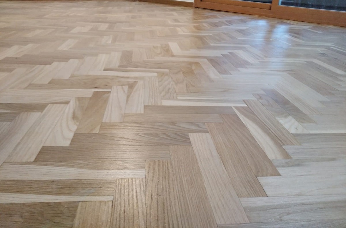 Jasny parkiet ułożony we wzór jodełki francuskiej, widoczne delikatne refleksy światła na polakierowanej powierzchni.