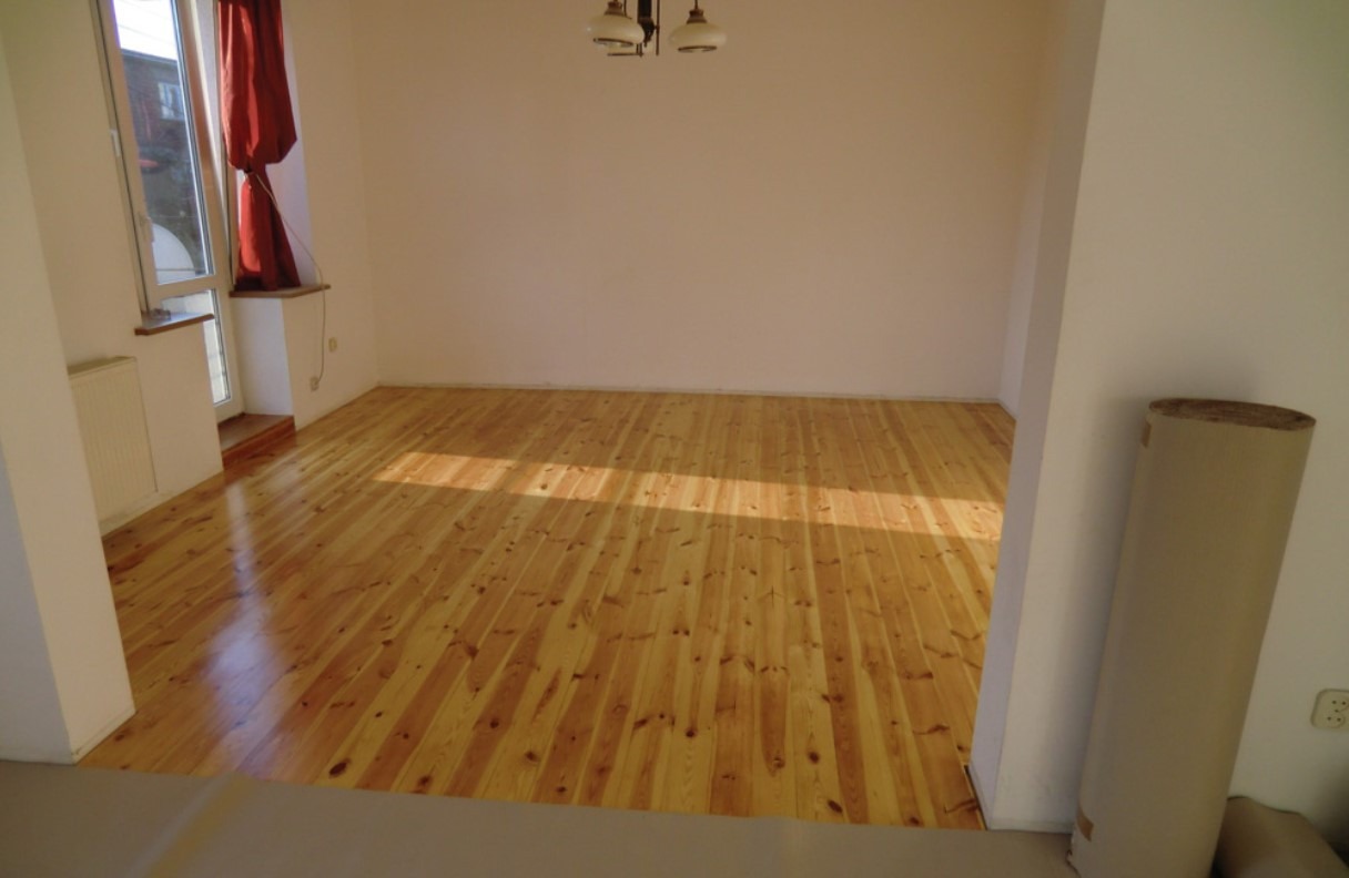 Wypolerowana drewniana podłoga z desek w pustym pokoju, widoczne promienie słoneczne i zwój materiału ochronnego przy ścianie.