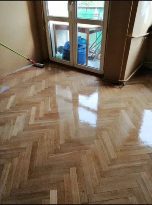 Świeżo polakierowany parkiet ułożony w jodełkę, odbijający światło z okna balkonowego, z widocznym mopem przy ścianie.