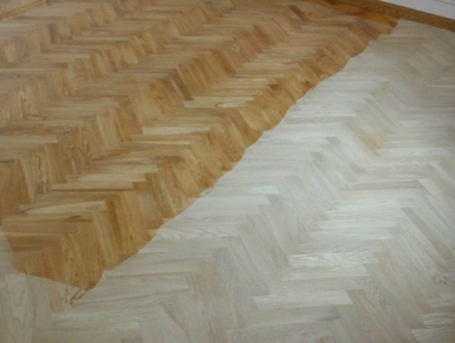 Fragment podłogi z parkietu ułożonego w jodełkę, częściowo po procesie lakierowania lub olejowania, z widocznym kontrastem między surowym, jasnym drewnem a zabezpieczoną, ciemniejszą powierzchnią.