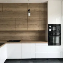 Nowoczesna kuchnia z białymi szafkami na wysoki połysk i drewnianymi panelami górnymi, czarna płyta indukcyjna i piekarnik w zabudowie, wisząca lampa bez klosza.